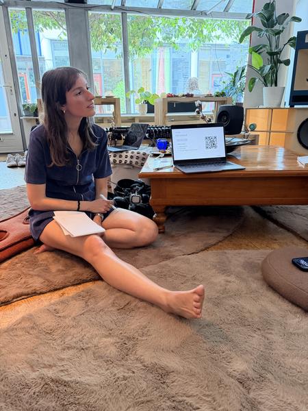 A person sits barefoot on a plush brown rug, looking thoughtfully towards a large window in an indoor space located at Koningsstraat, Nieuwmarktbuurt, Amsterdam